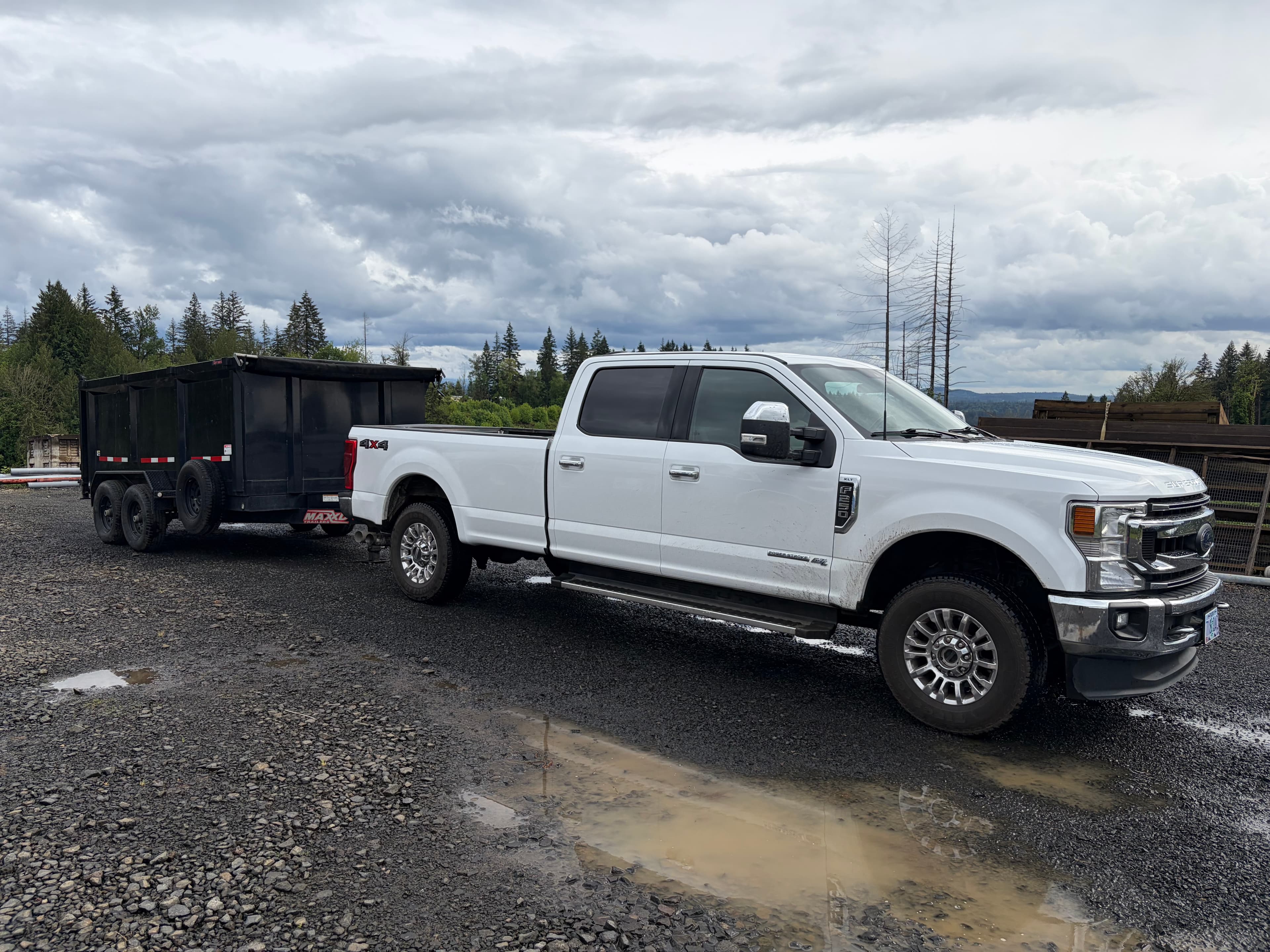 White Ford F-250 truck towing a black dump trailer on a wet gravel lot.