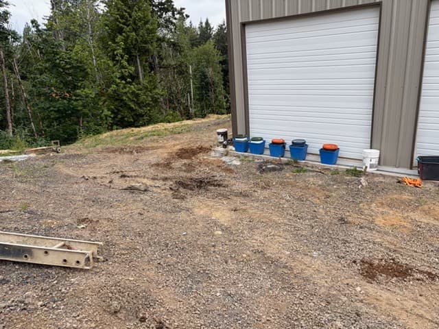 Blue bins lined up against a metal building with white garage doors near a forest.