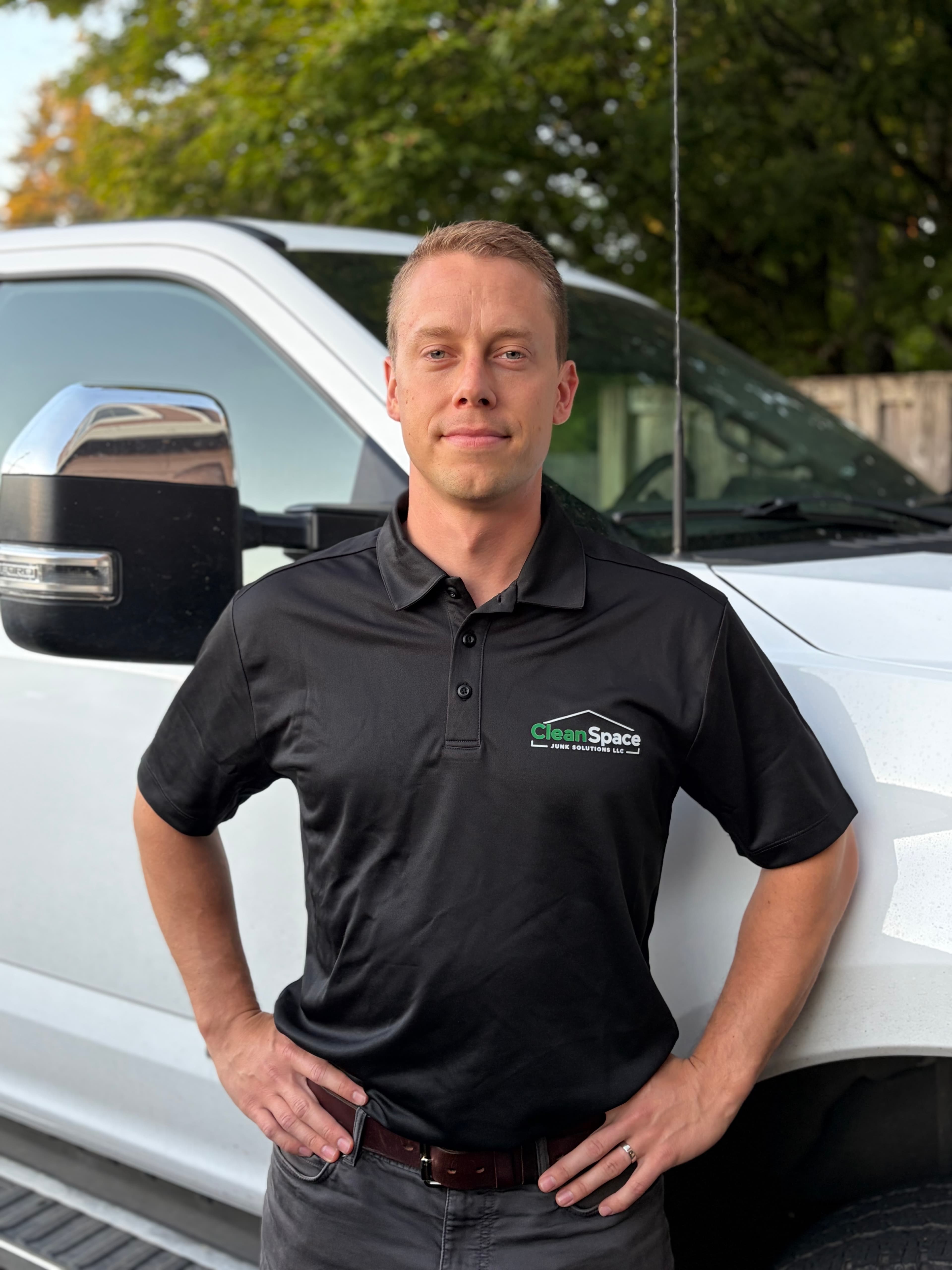 Man wearing a black CleanSpace Junk Solutions polo shirt posing by a white work truck.