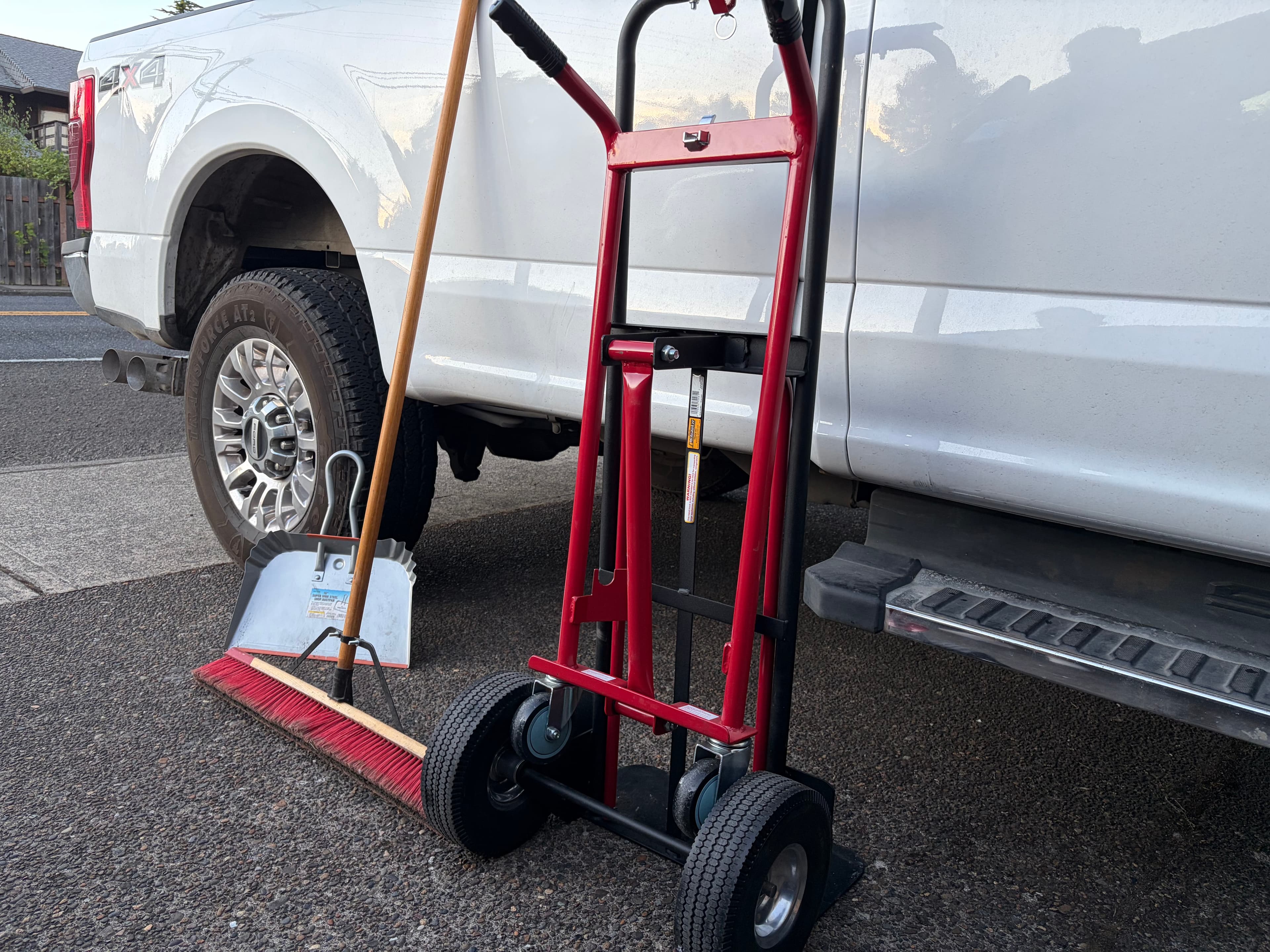 Red hand truck, push broom, and metal dustpan leaning against a white pickup truck.